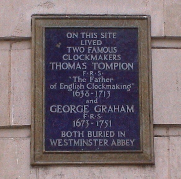 A blue plaque with white text. Read: "On this site lived two famous clockmakers Thomas Tompion F.R.S. The Father of English Clockmaking 1638-1713 and George Graham F.R.S. 1673-1751. Both bured at Westminster Abbey."